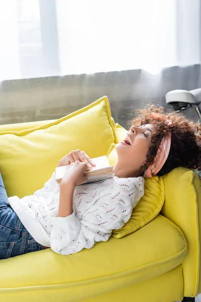 Excited young woman holding book while lying on sofa in living room — Stock Photo