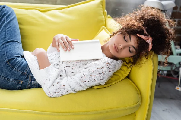 Кучерява молода жінка спить з книгою у вітальні — стокове фото