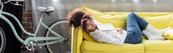 Horizontal crop of curly young woman reading book in living room — Stock Photo