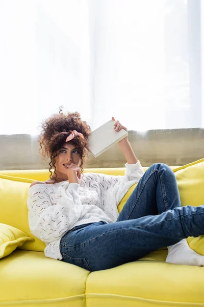 Nachdenkliche junge Frau hält Buch im Wohnzimmer — Stockfoto
