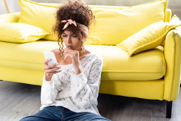 Беспокойная женщина смотрит на смартфон рядом с желтым диваном — стоковое фото