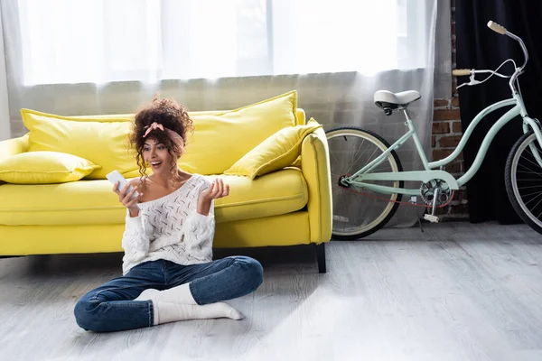 Curly woman looking at smartphone and laughing near yellow sofa — Stock Photo