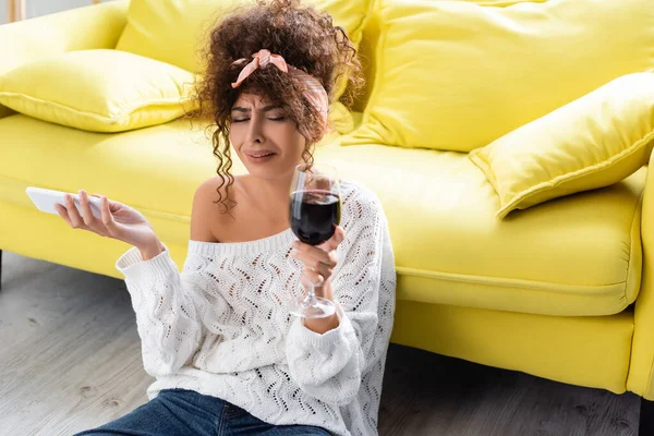 Traurige Frau hält Glas Wein und Smartphone in der Hand und weint im Wohnzimmer — Stockfoto