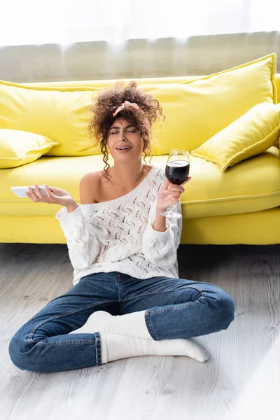 Mujer rizada sosteniendo copa de vino y teléfono inteligente mientras llora en la sala de estar - foto de stock