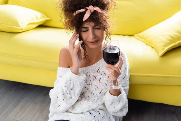 Curly woman holding glass of wine and talking on smartphone while crying in living room — Stock Photo