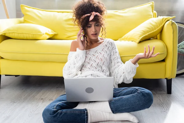 Dissatisfied freelancer sitting on floor with laptop and talking on smartphone — Stock Photo