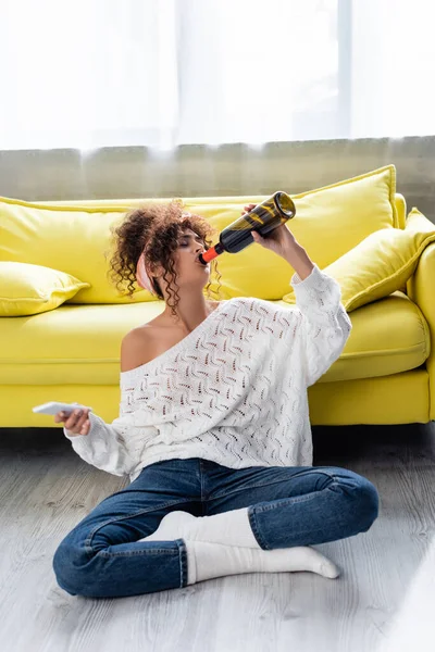 Curly woman drinking wine from bottle and holding smartphone — Stock Photo