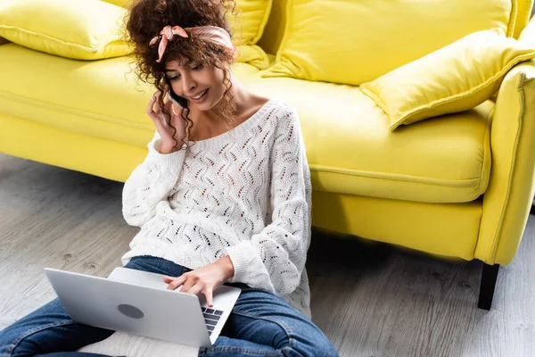 Alegre freelancer sentado en el suelo con ordenador portátil y hablando en el teléfono inteligente - foto de stock