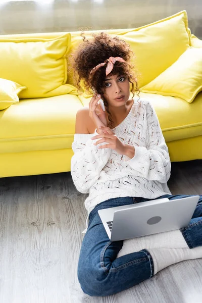 Curly freelancer sitting on floor with laptop and talking on smartphone — Stock Photo