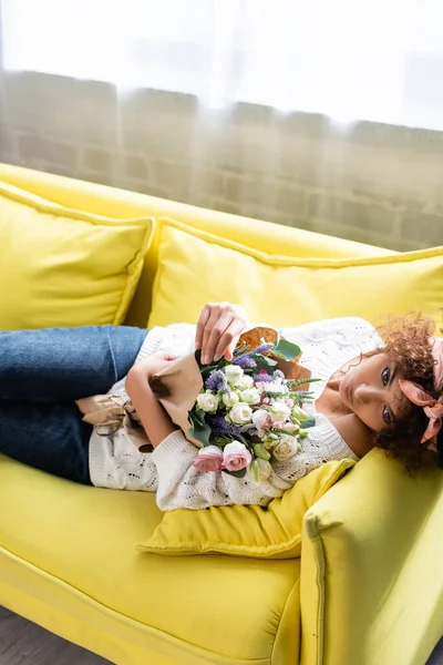 Молода кучерява жінка лежить на жовтому дивані з квітами і дивиться в сторону — стокове фото