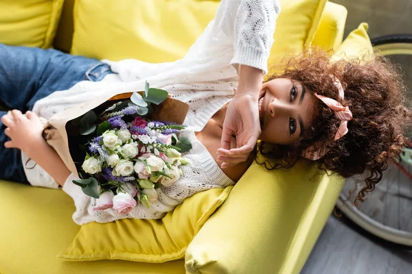 Vue de dessus de la femme bouclée avec des fleurs riant tout en étant couché sur le canapé — Photo de stock