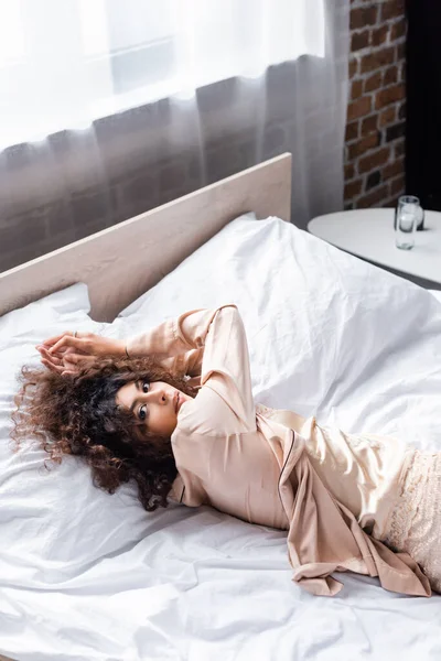 High angle view of curly and young woman in silk pajama lying on bed — Stock Photo