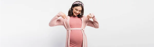 Horizontal concept of joyful pregnant woman pointing with fingers at belly on white — Stock Photo