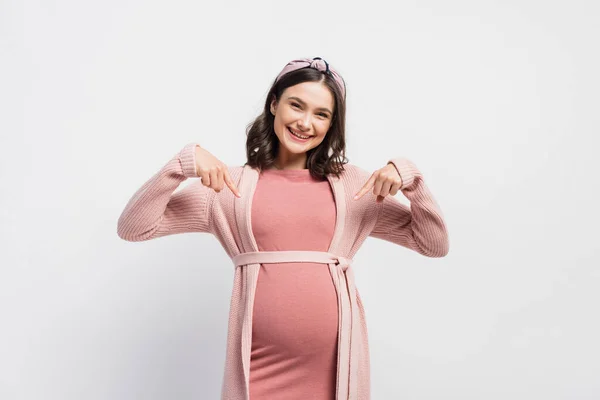 Joyful and pregnant woman pointing with fingers at belly on white — Stock Photo
