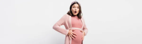 Panoramic shot of shocked and pregnant woman looking at camera while touching belly on white — Stock Photo