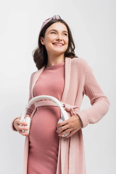 Mujer embarazada sosteniendo auriculares inalámbricos cerca del vientre aislado en blanco - foto de stock