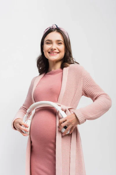 Mujer embarazada sosteniendo auriculares inalámbricos cerca del vientre aislado en blanco - foto de stock