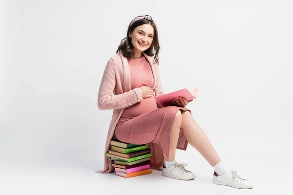 Mujer embarazada alegre sentada en los libros y tocando el vientre en blanco - foto de stock