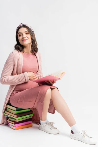 Pleased and pregnant woman sitting on books and touching belly on white — Stock Photo