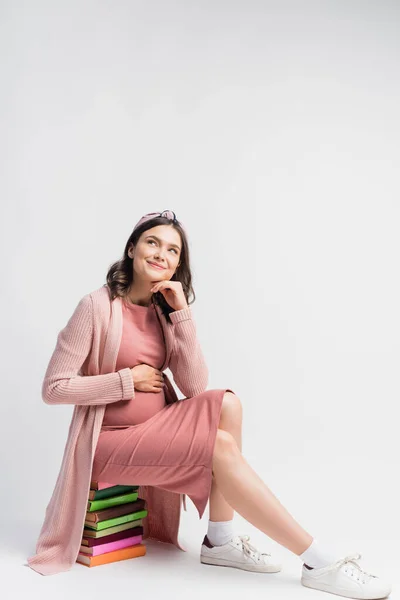Zufriedene und schwangere Frau, die auf Büchern sitzt und weiß wegsieht — Stockfoto