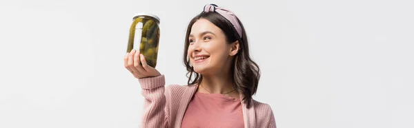 Panoramic shot of brunette woman in headband looking at jar with pickled cucumbers isolated on white — Stock Photo