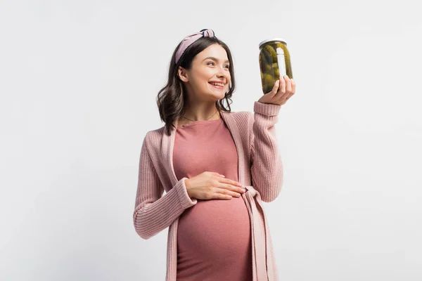 Joyful pregnant woman in headband looking at jar with pickled cucumbers isolated on white — Stock Photo