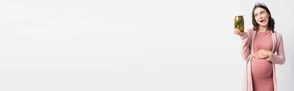 Панорамний урожай вагітної жінки, що стирчить язиком, дивлячись на банку з маринованими огірками, ізольовані на білому — стокове фото