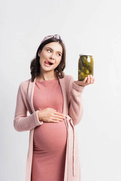 Femme enceinte qui sort la langue tout en regardant le pot avec des concombres marinés isolés sur blanc — Photo de stock