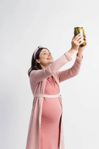 Heureux et femme enceinte tenant pot avec des concombres marinés au-dessus de la tête isolé sur blanc — Photo de stock