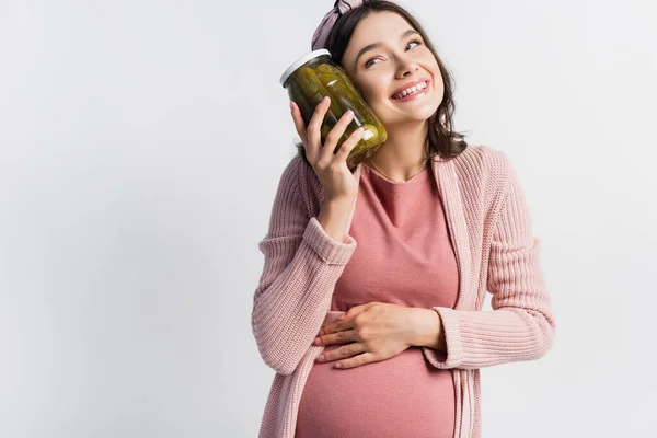 Mujer complacida y embarazada sosteniendo frasco con pepinos en escabeche aislados en blanco - foto de stock