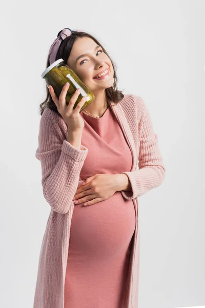 Довольная и беременная женщина держит банку с маринованными огурцами и смотрит в сторону изолированы на белом — стоковое фото