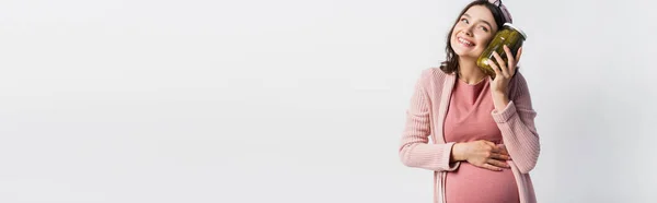 Panoramic crop of pleased and pregnant woman holding jar with pickled cucumbers and looking away isolated on white — Stock Photo