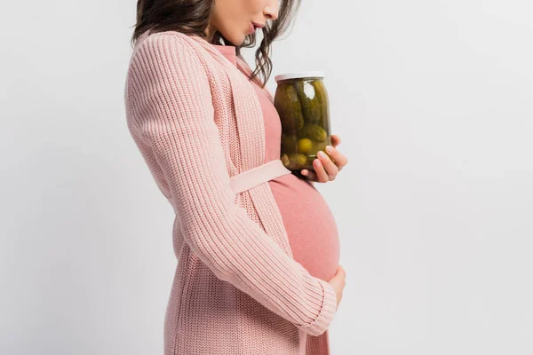 Vista parcial de la mujer sorprendida y embarazada sosteniendo frasco con pepinos en escabeche aislados en blanco - foto de stock