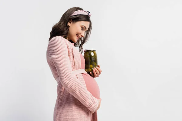 Visão lateral da mulher satisfeita e grávida segurando frasco com pepinos em conserva isolados no branco — Fotografia de Stock
