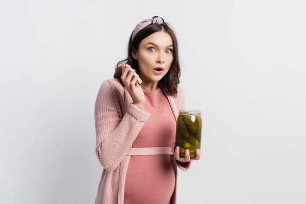 Surprised and pregnant woman holding jar with pickled cucumbers on white — Stock Photo