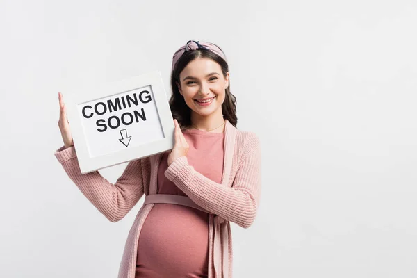 Zufriedene und schwangere Frau hält Tafel mit bald erscheinendem Schriftzug isoliert auf weißem Grund — Stockfoto