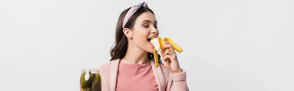Panoramic shot of woman eating banana and holding jar with canned cucumbers isolated on white — Stock Photo