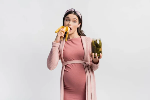 Donna incinta mangiare banana e guardando vaso con cetrioli in scatola isolato su bianco — Foto stock