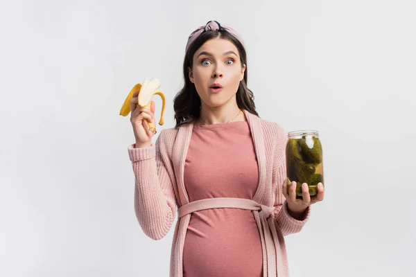 Pregnant woman eating banana while holding jar with canned cucumbers isolated on white — Stock Photo