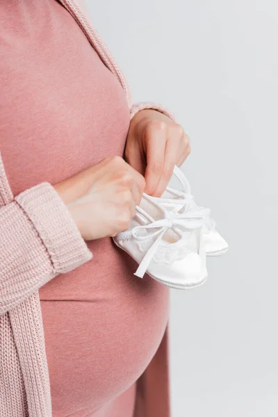 Vista recortada de la mujer embarazada sosteniendo pequeñas botas de bebé aisladas en blanco - foto de stock