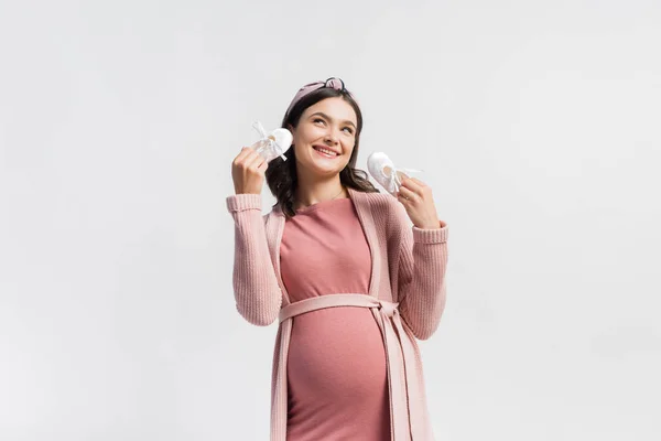 Mujer alegre y embarazada sosteniendo pequeñas botas y mirando hacia arriba aislado en blanco - foto de stock