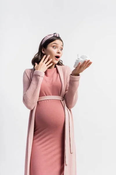 Shocked and pregnant woman looking at tiny boots isolated on white — Stock Photo