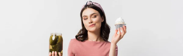 Panoramic shot of woman choosing between cupcake and jar with pickled cucumbers isolated on white — Stock Photo