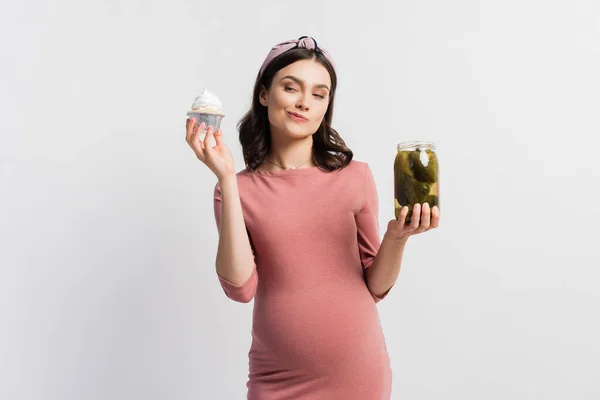 Pregnant woman choosing between cupcake and jar with pickled cucumbers isolated on white — Stock Photo