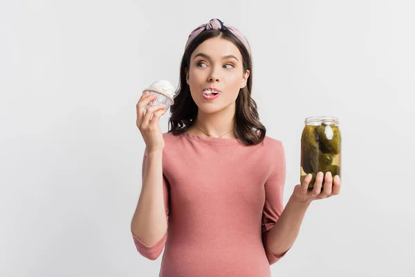 Mujer embarazada comiendo magdalena y sosteniendo frasco con pepinos en escabeche aislados en blanco - foto de stock