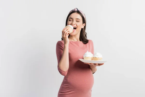 Joyful pregnant woman holding plate while eating cupcake isolated on white — Stock Photo