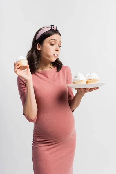 Joven embarazada sosteniendo plato mientras come cupcake aislado en blanco - foto de stock