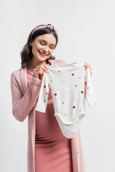 Joyful pregnant woman in headband holding baby romper isolated on white — Stock Photo