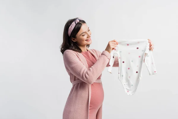 Joyful pregnant woman in headband looking at baby romper isolated on white — Stock Photo