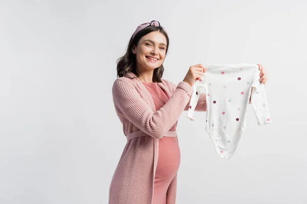 Pleased pregnant woman in headband holding baby romper isolated on white — Stock Photo